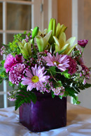 A glass purple square vase filled with lavender daisies, lavender poms, white lilies, fuchsia carnations