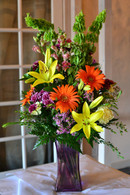 Two thumbs up for mom, a cheerful bright bouquet, filled with vibrant yellow lilies, green bells of Ireland, Orange gerber daisies, purple poms, lavender wax flower put together in a purple square tall vase
