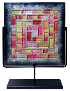 A square fused glass panel featuring red and pink mosaic-like tiles surrounded by black.  Stand included