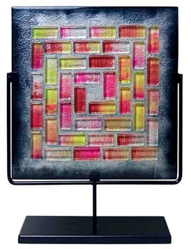 A square fused glass panel featuring red and pink mosaic-like tiles surrounded by black.  Stand included