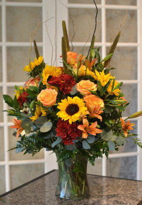 A large show stopper! A clear vase filled with cattails, sunflowers, curly willow, mums, lilies & roses