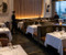 An elegant, high-end fine dining restaurant that masterfully blends mid-century modern sophistication with contemporary luxury. The space utilizes a rich combination of natural textures—Spirit Birch wood paneling, plush velvet, smooth leather, and dark stone—to create an intimate, upscale atmosphere with a sweeping urban backdrop.