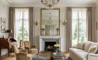 The slender white marble mantel The Versailles stands proudly on a central wall breast, framing a deep, gray-toned firebox equipped with traditional brass andirons, logs, and a low brass hearth fender. It is topped with a classical marble bust that anchors the mantel design.