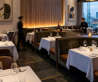 An elegant, high-end fine dining restaurant that masterfully blends mid-century modern sophistication with contemporary luxury. The space utilizes a rich combination of natural textures—Spirit Birch wood paneling, plush velvet, smooth leather, and dark stone—to create an intimate, upscale atmosphere with a sweeping urban backdrop.