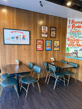 The MCM Spirit Birch wood-paneled walls provide a backdrop for colorful retro-style posters and framed artwork, while a large, hand-painted typography mural adds a playful, nostalgic character to the space. Dark wood-look flooring complements the paneling, and teal-blue upholstered chairs with silver metal frames introduce a contrasting pop of color.