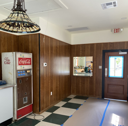 A nostalgic, retro-inspired commercial space, community breakroom, or recreation hall that captures a mid-to-late 20th-century Americana vibe. The room is currently undergoing partial renovation or finishing work, pairing vintage elements like wood paneling and a checkered floor with traditional fixtures.
